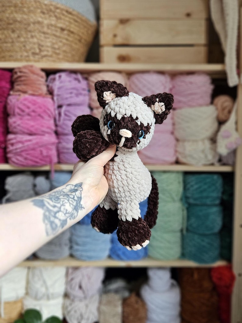 May include: A handmade crochet cat plushie in brown, white, and light blue. The cat has dark brown paws, ears, and facial markings, with light blue eyes and a white muzzle. The plushie is held in front of a shelf filled with yarn.