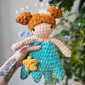 May include: A crocheted fairy doll with brown hair, blue wings, and a teal dress. The doll is holding a yellow flower.