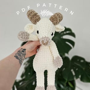 Może przedstawiać: Pluszowa zabawka w kształcie kozy, wykonana na szydełku, z brązowymi rogami i biało-brązowym futerkiem. Zabawka jest trzymana przez rękę z tatuażem.