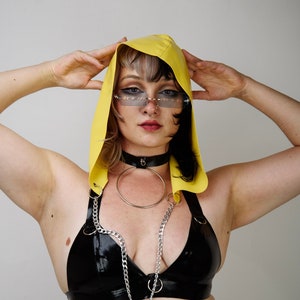 Puede incluir: Una persona lleva una capucha amarilla, un sujetador de látex negro con una cadena plateada, un collar de gargantilla plateado con un anillo y una falda de látex negra.