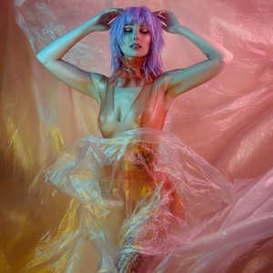 May include: A person with purple hair poses in a sheer, iridescent fabric. The fabric is draped over the person's body, creating a flowing, ethereal effect. The person's face is visible, and they are looking down.