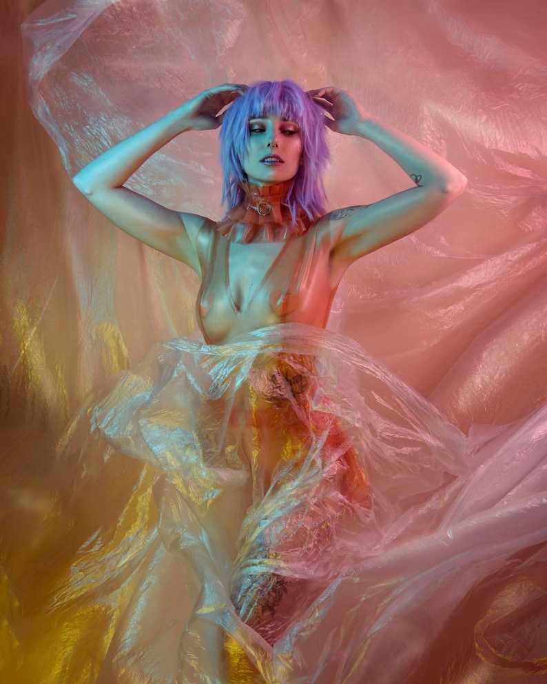 May include: A person with purple hair poses in a sheer, iridescent fabric. The fabric is draped over the person's body, creating a flowing, ethereal effect. The person's face is visible, and they are looking down.