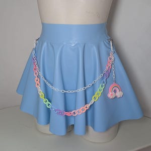 May include: A light blue skirt with a pleated design. A decorative chain with pastel-colored links and a rainbow charm hangs from the skirt. The skirt is displayed on a mannequin.