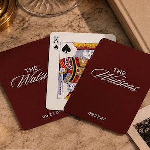 Puede incluir: Cartas de juego color burdeos con "The Watsons" en escritura elegante y la fecha "09.27.27". Se ve una carta de Rey de Picas. Un anillo dorado con una "W" negra está cerca de un libro titulado "A Demeure Beautiful".