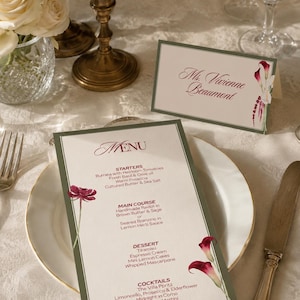 May include: Elegant table setting with a menu and place card. The menu features a floral design and a list of dishes. The place card reads "Ms. Vivienne Beaumont". A vase of white roses and gold candle holders are also visible.