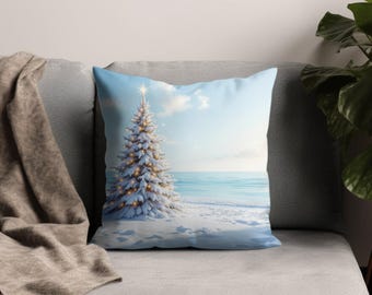 Puede incluir: Un cojín decorativo con una escena de playa invernal. Un árbol de Navidad cubierto de nieve con luces doradas se alza en una orilla nevada, con un océano y un cielo azul brillante en el fondo. El cojín está sobre un sofá gris.