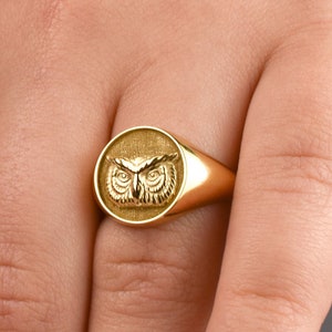 May include: A gold signet ring featuring a circular face with an embossed owl design. The owl is depicted with detailed feathers and prominent eyes centered on the flat, round surface of the ring. The band is smooth and polished gold.