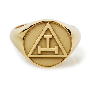 Puede incluir: Un anillo de sello dorado con una cara circular. La cara presenta un triángulo con una línea horizontal y una línea vertical en el centro. El anillo está sobre un fondo blanco.