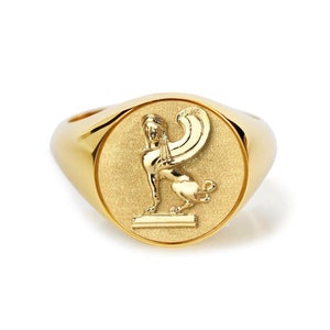 May include: A gold signet ring featuring a raised design of a winged lion on a square base.