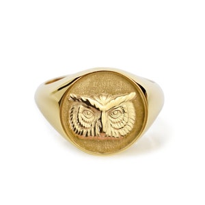 May include: A gold signet ring featuring a circular face with a detailed, raised relief of an owl head. The owl design is centered on a textured, matte-finish background, while the band and the outer edge of the ring face have a polished, smooth, and reflective gold surface.
