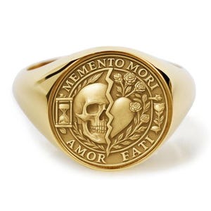 May include: A gold-colored signet ring with a detailed design. The ring features a circular emblem with a skull, a heart, and the phrases "Memento Mori" and "Amor Fati". The ring has a polished finish and a classic, elegant style.