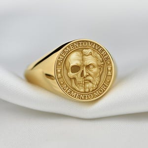 May include: A gold signet ring with a detailed design. The ring features a split face with a skull on one side and a bearded man on the other, encircled by the phrases "MEMENTO MORI" and "MEMENTO VIVERE".