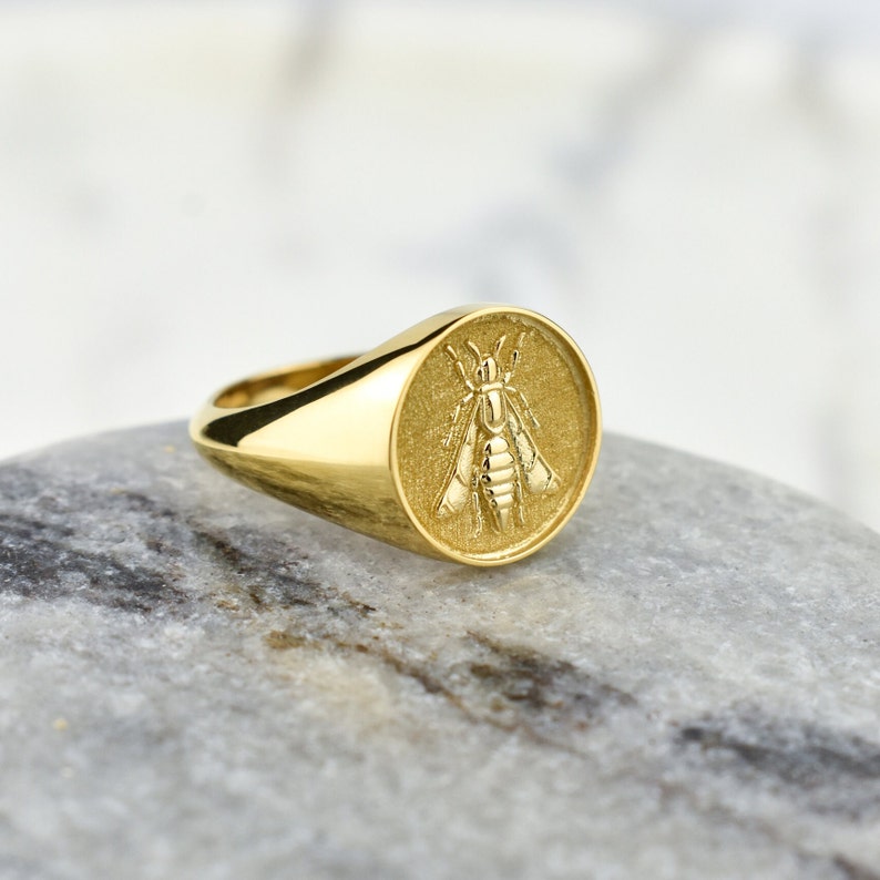 Puede incluir: Un anillo de sello en tono dorado con una cara circular que presenta un emblema de abeja en relieve y detallado. El anillo tiene una banda lisa y pulida, y un fondo texturizado con acabado mate detr&aacute;s del motivo de la abeja.