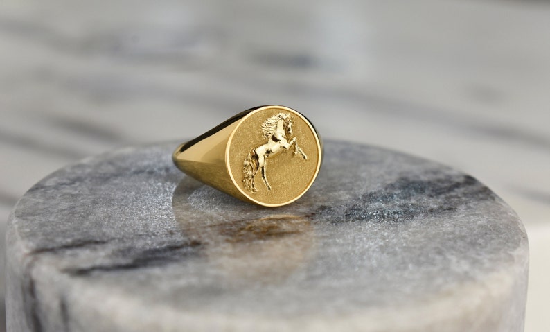 Puede incluir: Un anillo de sello de oro con un dise&ntilde;o en relieve de un caballo en la cara del anillo.