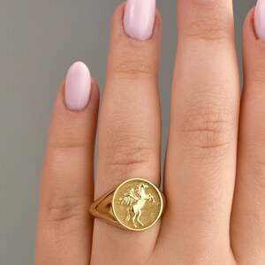 May include: A gold signet ring featuring a raised design of a winged horse. The ring is worn on a finger with pink nail polish.