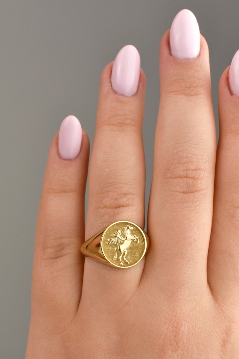 May include: A gold signet ring featuring a raised design of a winged horse. The ring is worn on a finger with pink nail polish.
