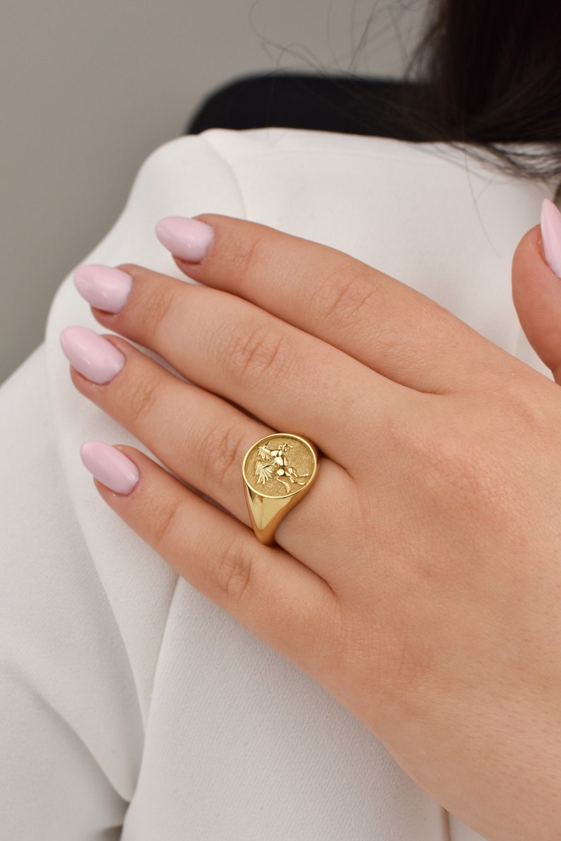 May include: A gold signet ring featuring a raised design of a mythical creature. The ring is worn on a finger with light pink nail polish.