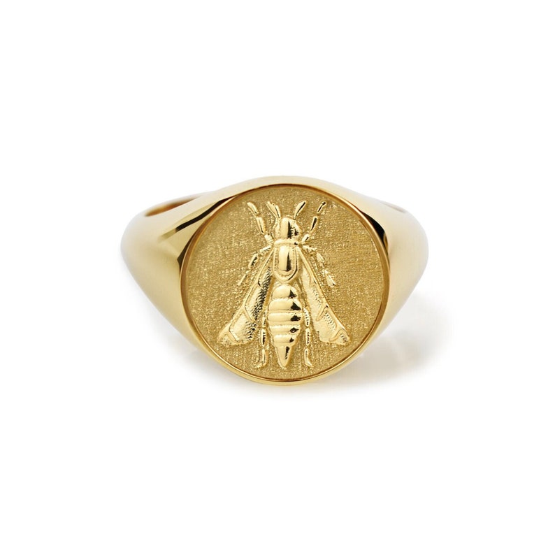 Puede incluir: Un anillo de sello dorado con una cara circular que presenta un emblema de abeja detallado en relieve. El anillo tiene una banda lisa y pulida, con un fondo texturizado detr&aacute;s del motivo de la abeja, creando un contraste entre superficies.