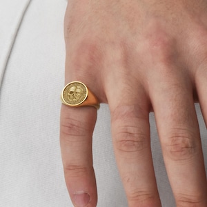 May include: A gold signet ring featuring a skull design. The ring is worn on a finger, showcasing its polished surface and intricate details. The ring's design includes the words "MEMENTO MORI" around the skull.