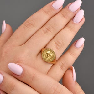 May include: A gold-colored signet ring featuring a skull design with the words "Memento Mori" engraved around the edge. The ring is shown on a hand with light pink nail polish.