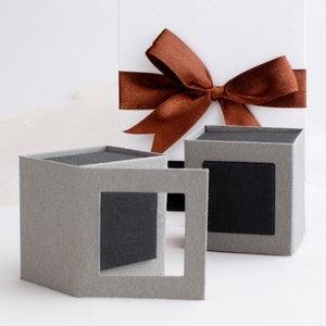 May include: Two grey gift boxes with black inserts. The boxes are open and the inserts are visible. The boxes are on a white surface. A white gift box with a brown ribbon is in the background.