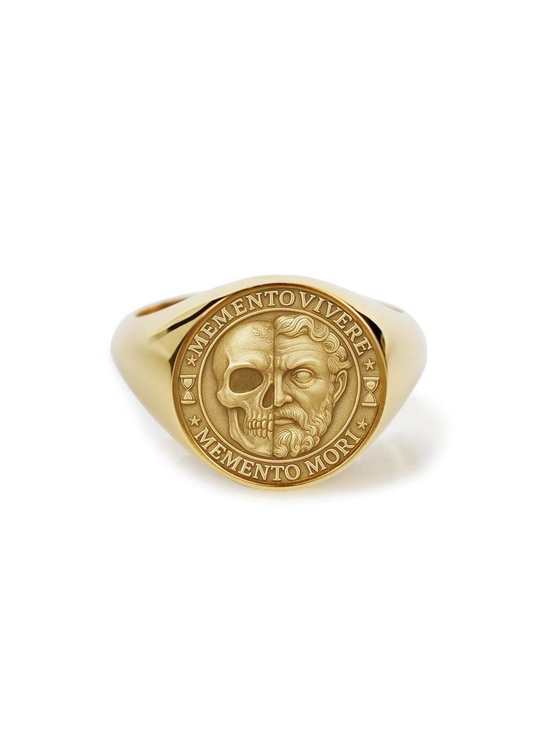 May include: A gold signet ring featuring a detailed design. The ring's face is split, with a skull on one side and a bearded face on the other. The words "MEMENTO MORI" and "MEMENTO VIVERE" are inscribed around the design.