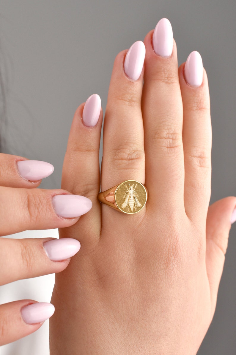 Puede incluir: Un anillo de sello de oro con una cara circular que presenta un dise&ntilde;o de abeja grabado. El anillo tiene un acabado pulido y una banda lisa y redondeada que se estrecha hacia la parte posterior, mostrando un insecto detallado en el centro.