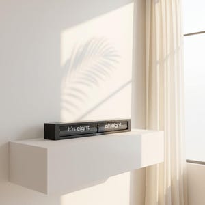 May include: A black digital clock displaying "It's eight oh eight" on a white shelf against a white wall. The clock has a rectangular shape and a modern design. The text is in a clean, sans-serif font.