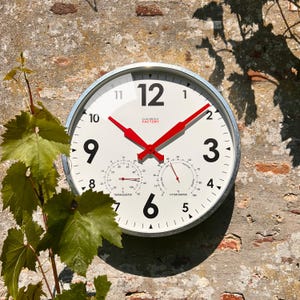 May include: A white outdoor clock with a silver frame and red hands. The clock face has black numbers and includes a thermometer and hygrometer. The clock face also includes the text "CLOVONGLA FACTORY".