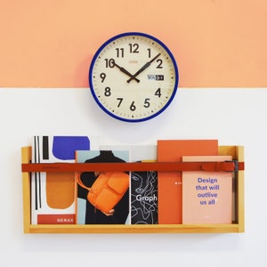 May include: A blue-rimmed wall clock with black hands and large numerals hangs above a wooden shelf. The shelf holds books and a bag, secured by a red leather strap. The text "Design that will outlive us all" is visible on a book.