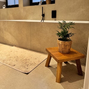 Peut inclure: Un tabouret en bois avec un pot de plante en osier tissé sur le dessus. Le tabouret se trouve dans une salle de bain avec un sol en carrelage beige et une baignoire blanche.