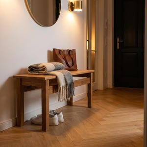 Op de afbeelding: Een houten bank met een opgevouwen deken en een leren draagtas. De bank staat in een hal met een parketvloer, een ronde spiegel en een wandlamp. Een paar witte sneakers staan onder de bank.