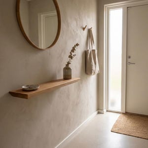 Op de afbeelding: Een minimalistische entree met een houten plank, een ronde spiegel en een beige draagtas aan de muur. Een kleine vaas met groen staat op de plank. De deur en muren zijn geschilderd in neutrale tinten.