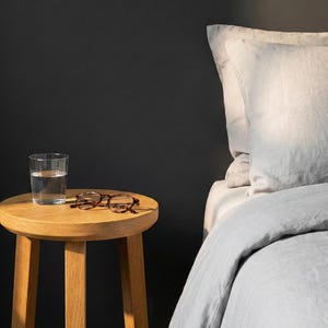 Könnte beinhalten: Ein Glas Wasser und eine Brille mit Schildpattmuster liegen auf einem kleinen, runden Beistelltisch aus Holz. Im Hintergrund befindet sich ein Bett mit einer hellgrauen Bettdecke und Kissen. Der Raum hat eine dunkelgraue Wand.