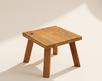 May include: A small, square wooden stool crafted from light brown oak with a visible natural wood grain pattern. The stool features four sturdy, angled legs and a flat top surface with a small, dark knot detail near the left corner edge.