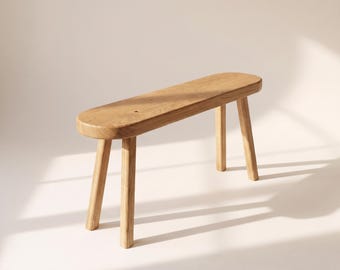May include: A light brown wooden bench featuring a smooth, elongated oval seat with rounded edges and four sturdy, slightly angled legs. The natural wood grain is visible across the surface, showcasing a warm, light-toned finish throughout.