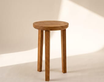May include: A small wooden stool featuring a circular top and three straight, sturdy legs. The stool is crafted from light brown wood with a visible natural grain pattern, showcasing a simple, functional design with a smooth, polished finish.