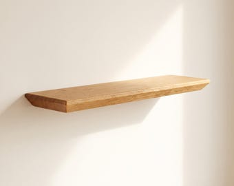 May include: A rectangular floating wall shelf made of light brown wood with a visible natural grain pattern. The shelf features a smooth, flat top surface and angled, beveled edges on the front and sides, creating a clean, solid appearance.