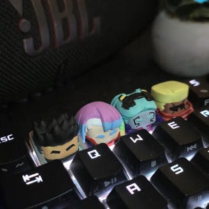 Puede incluir: Cuatro teclas artesanales personalizadas con forma de cabezas de personajes estilizadas están colocadas en un teclado mecánico negro. Las teclas presentan colores de cabello distintos, incluyendo negro, rosa y azul, turquesa y amarillo.