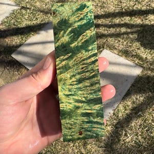 Könnte beinhalten: Ein rechteckiges Holzstück mit einem lebendigen grün-goldenen Wirbelmuster. Die Holzmaserung ist sichtbar und erzeugt eine einzigartige Textur. Das Stück wird in einer Hand gehalten, um seine Größe und Details zu zeigen. Das Holz scheint ein dekoratives Element zu sein.