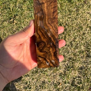 Op de afbeelding: Een rechthoekig stuk donkerbruin hout met een uniek nerfpatroon, vastgehouden in een hand. Het hout heeft een gepolijste, glanzende afwerking en de nerf creëert een wervelend, gestructureerd uiterlijk. Het hout is ongeveer 13 cm lang.