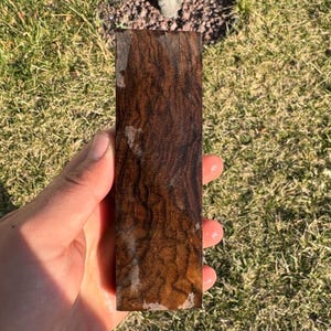 Könnte beinhalten: Ein rechteckiges Holzstück, dunkelbraun mit detaillierten Maserungsmustern. Das Holz hat ein poliertes Aussehen mit helleren Streifen und Farbtonvariationen. Das Holz wird in einer Hand gehalten, mit einem grasbewachsenen Hintergrund.