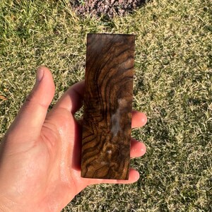 Könnte beinhalten: Ein rechteckiges Holzstück mit einer satten, dunkelbraunen Farbe und einem einzigartigen, wirbelnden Maserungsmuster. Das Holz hat eine polierte, glänzende Oberfläche und wird in einer Hand vor grünem Gras gehalten.