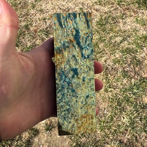 Könnte beinhalten: Ein rechteckiger Holzblock mit einem einzigartigen, wirbelnden Muster aus blauen, goldenen und braunen Farbtönen. Die Holzmaserung ist sichtbar und erzeugt ein strukturiertes Aussehen. Der Block wird in einer Hand gehalten, um seine Größe und Form zu zeigen.