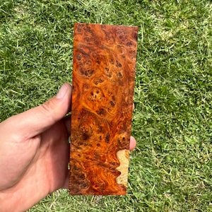 Könnte beinhalten: Ein rechteckiges Holzstück mit einem einzigartigen Maserungsmuster. Das Holz weist eine Mischung aus rotbraunen und goldenen Farbtönen mit komplexen Wirbelmustern auf. Das Holz wird vor einem Hintergrund aus grünem Gras gehalten.