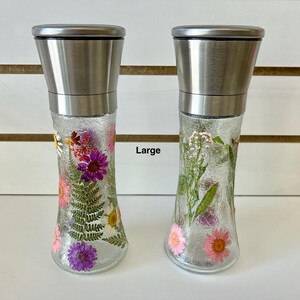 May include: Two clear glass spice grinders with stainless steel tops. The grinders are filled with dried flowers in shades of pink, purple, and white. The word "Large" is printed on the image.