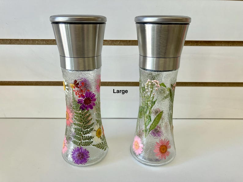 May include: Two clear glass spice grinders with stainless steel tops. The grinders are filled with dried flowers in shades of pink, purple, and white. The word "Large" is printed on the image.