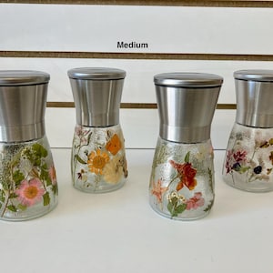 May include: Four glass spice grinders with stainless steel tops. The clear glass bases are decorated with pressed flowers in various colors, including pink, orange, and red. The word "Medium" is above the grinders.