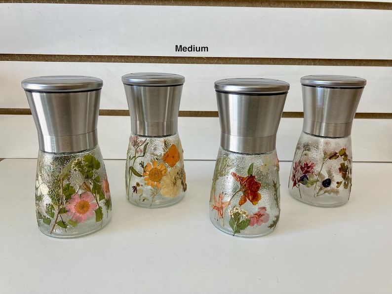 May include: Four glass spice grinders with stainless steel tops. The clear glass bases are decorated with pressed flowers in various colors, including pink, orange, and red. The word "Medium" is above the grinders.