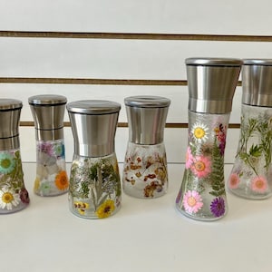 May include: Six glass spice grinders with stainless steel tops. The clear glass bodies are decorated with pressed flowers in various colors, including yellow, pink, and purple. The grinders are arranged on a white surface.